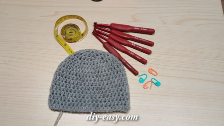 Gray crochet hat in progress with crochet hooks, measuring tape, and stitch markers on a wooden surface