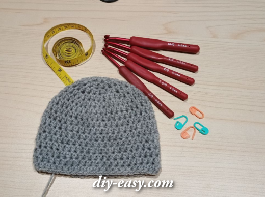 Gray crochet hat in progress with crochet hooks, measuring tape, and stitch markers on a wooden surface